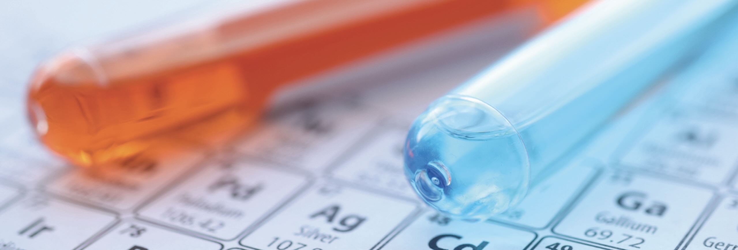 A photo of two test tubes, one containing blue liquid and one containing orange liquid, laying on a portion of the Periodic Table.